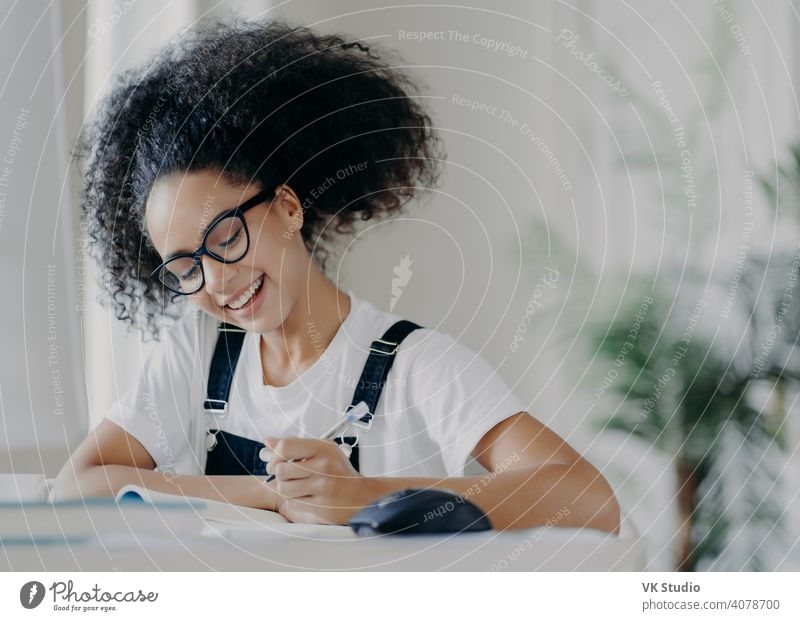 Glückliche afroamerikanische Studentin schreibt Informationen auf, bereitet sich auf Prüfungen vor, sitzt im Coworking Space, hat lockiges dunkles Haar, trägt eine optische Brille, weißes T-Shirt und Latzhose, studiert in einem geräumigen Zimmer