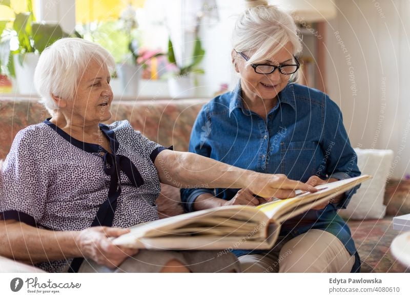Ältere Frau und ihre erwachsene Tochter betrachten gemeinsam ein Fotoalbum auf der Couch im Wohnzimmer Sitzen Gedächtnis Erinnerungen Nostalgie Historie Fotos