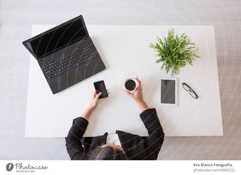Draufsicht auf eine junge Geschäftsfrau, die im Büro mit Handy arbeitet und eine Tasse Kaffee hält. Tagsüber. Lebensstile. Innenbereich Frau Computer Laptop