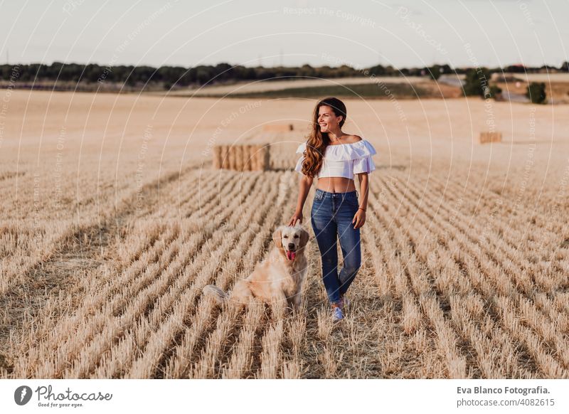 Junge schöne Frau, die mit ihrem Golden Retriever Hund auf einem gelben Feld bei Sonnenuntergang spazieren geht. Natur und Lebensstil im Freien Glanz Erholung