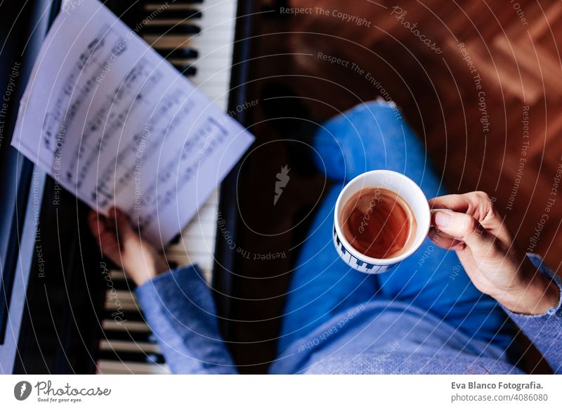 junge unerkennbare Frau, die eine Tasse Kaffee hält und Klavier spielt, indem sie ein Notenblatt liest. Musik-Konzept im Innenbereich. Ansicht von oben Stil