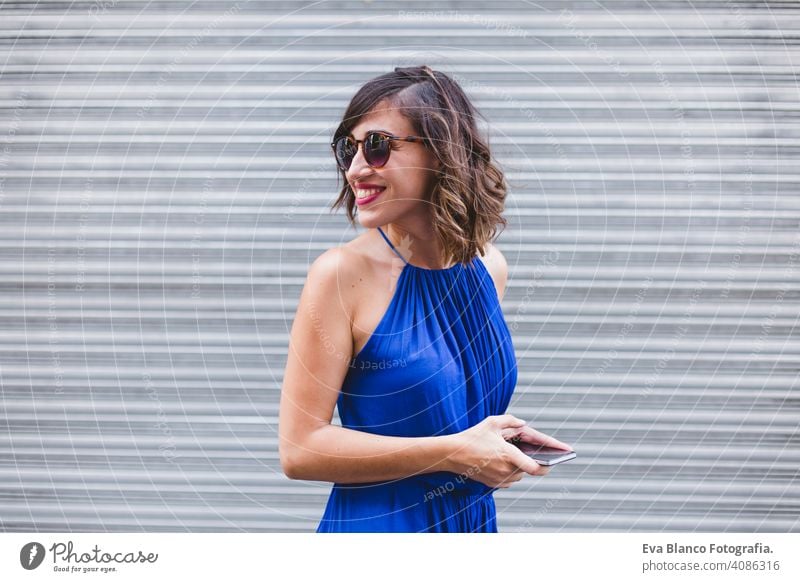 junge schöne Frau mit Handy und lächelnd. Sie ist auf der Straße, trägt ein lässiges blaues Kleid, Sonnenbrille und lächelnd. LIfestyle im Freien, modernes Leben und Technologie