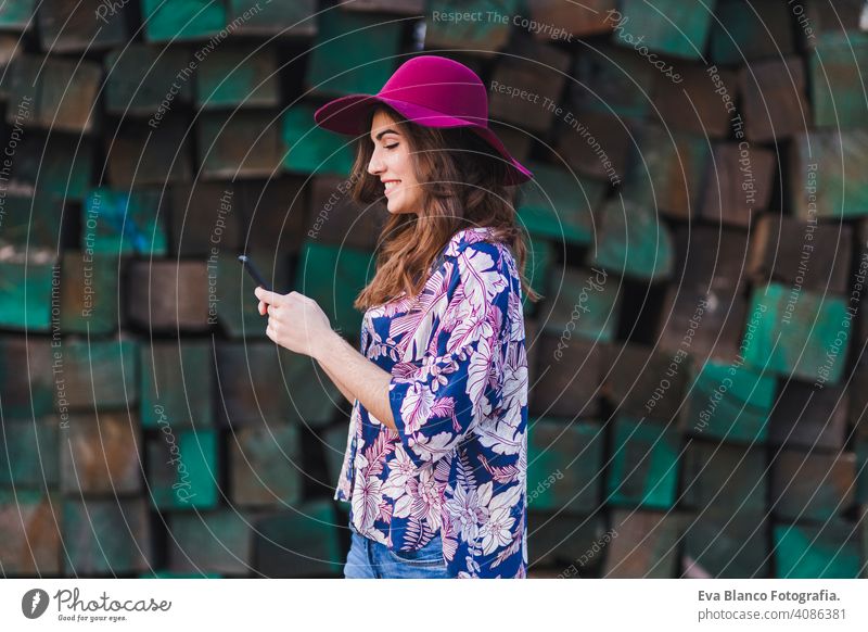 Porträt einer jungen schönen Frau, die legere Kleidung und einen modernen Hut trägt und ihr Mobiltelefon benutzt. Sie steht über grünen Holzblöcken Hintergrund und lächelnd. Lebensstil im Freien.