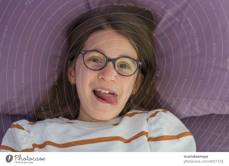 Nahaufnahme eines kleinen kaukasischen Mädchens mit Brille, das auf dem Bett liegt und die Zunge herausstreckt Sehvermögen horizontal Unschuld unschuldig lachen