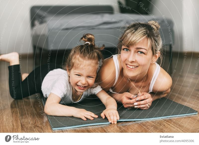 Eine schöne junge Frau und ein bezauberndes kleines Mädchen schauen in die Kamera und lächeln, während sie auf einer Yogamatte im Schlafzimmer liegen Fitness