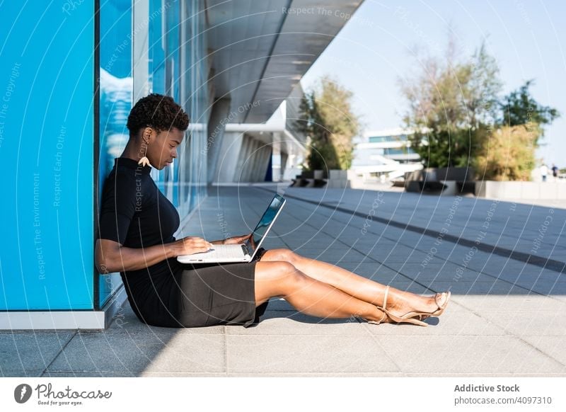 Seriöse Frau surft am Laptop in der Nähe eines modernen Gebäudes sich[Akk] entspannen konzentriert ernst benutzend Surfen Afroamerikaner elegant Straße Kleid