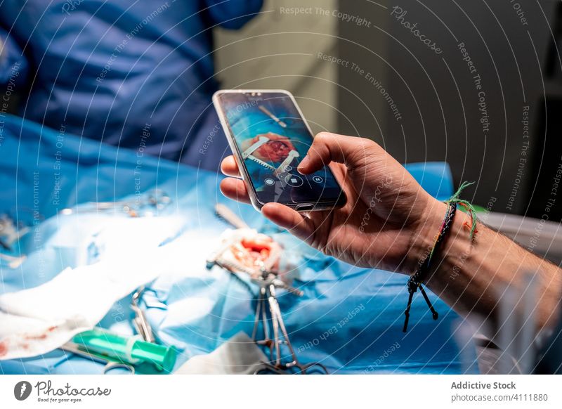 Mann fotografiert mit Smartphone, während medizinisches Personal Patienten in einer modernen Klinik operiert Chirurg Chirurgie Krankenhaus Sanitäter Hand Handy