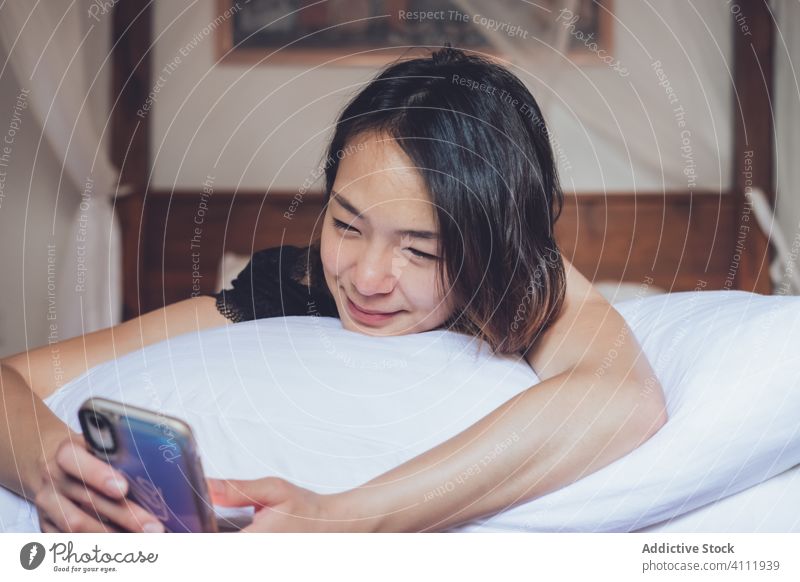 Glückliche asiatische Frau benutzt Smartphone im Bett benutzend Lächeln ethnisch Kopfkissen heimwärts gemütlich sich[Akk] entspannen ruhen Schlafzimmer jung