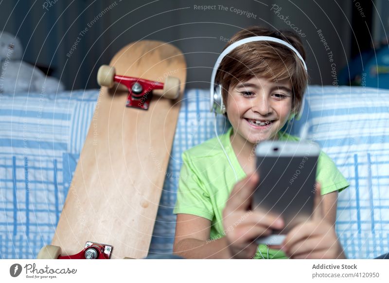 Fröhliches Kind mit Kopfhörern, das zu Hause sein Smartphone benutzt Apparatur heimwärts heiter Junge benutzend Lachen Talkrunde Gerät online zuhören Internet