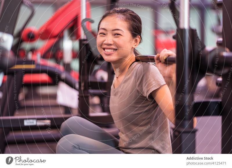 Positive asiatische Sportlerin lehnt sich in moderner Turnhalle an Langhantel und schaut in die Kamera Frau Curl-Hantel ruhen Training Fitnessstudio Maschine