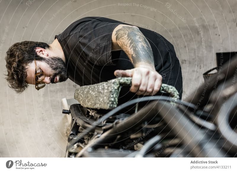 Beschäftigter männlicher Mechaniker bei der Reparatur eines Motorradsitzes in einer Werkstatt Biker Garage Mann fixieren messen Sitz Verkehr beschäftigt Tattoo