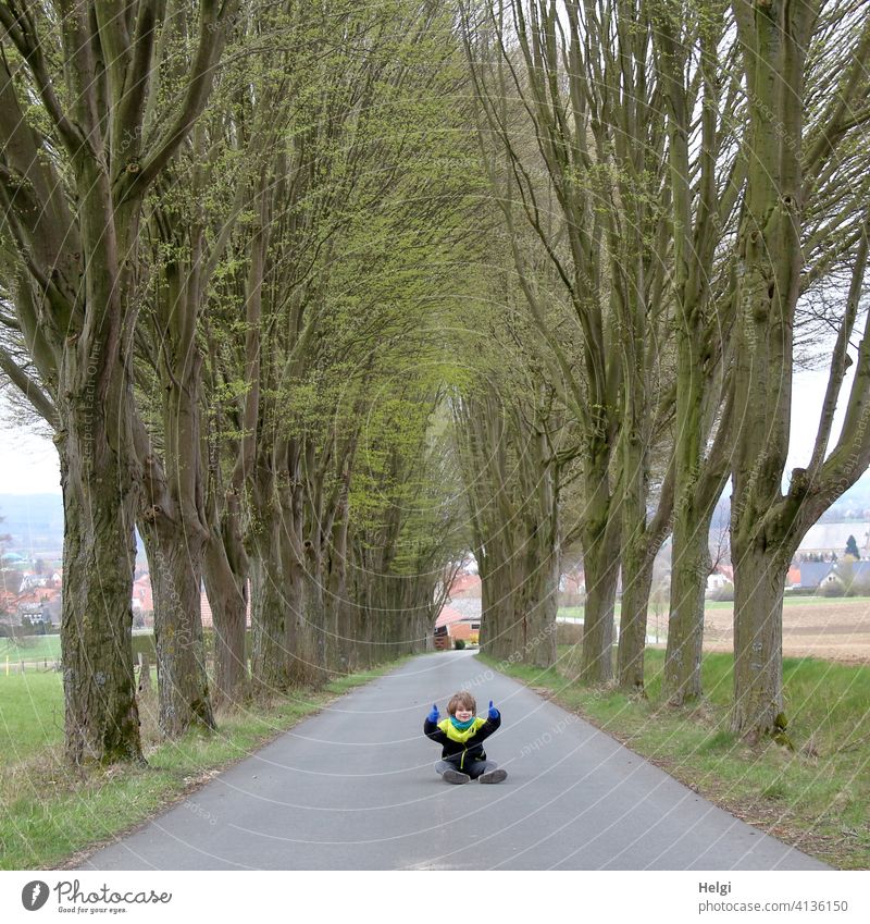 Kind sitzt mitten auf der unbefahrenen Straße einer Allee und hält die Daumen hoch Mensch Schulkind Asphalt sitzen Frühling Baum Feld Straßenrand Baumreihe