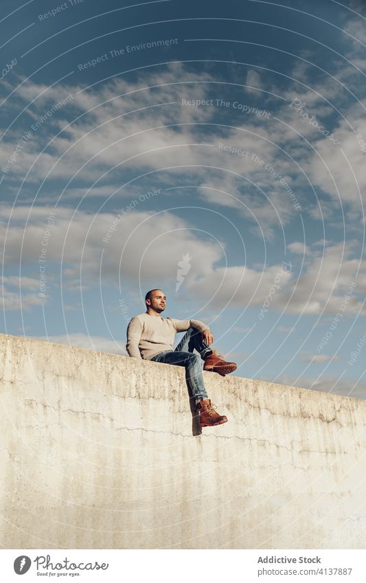 Hipster Mann sitzt auf Betonwand besinnlich Stil trendy modern Streetstyle nachdenklich nachdenken Zaun Wand jung unrasiert Typ Jeanshose sitzen lässig Himmel
