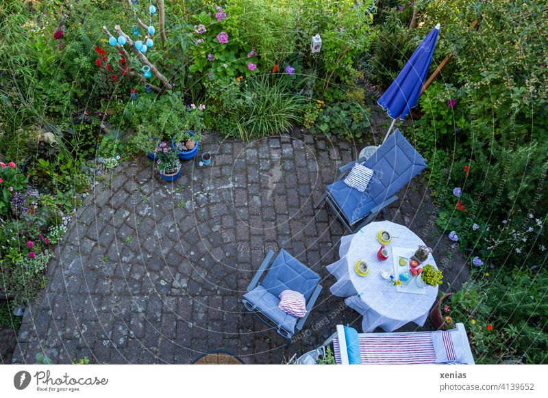 Vogelperspektive im Sommer: Zwei gelbe Tassen Kaffee auf dem Tisch, Stühle mit gemütlichen Kissen und Sonnenschirm stehen auf der Terrasse bereit. Diese ist umgeben von grünen Pflanzen mit bunten Blüten. Wo sind die Kaffeetrinker hin?