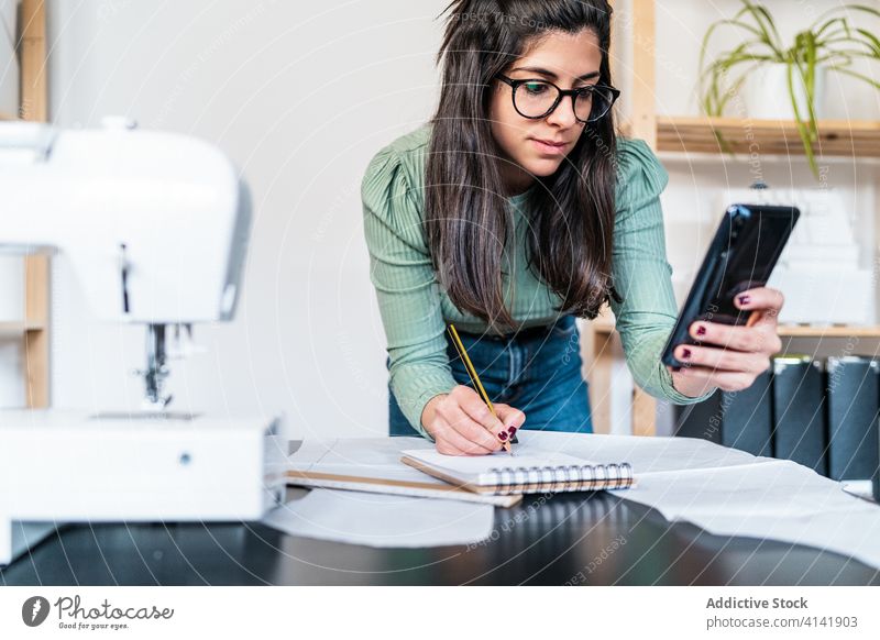 Ethnische Schneiderin, die in der Werkstatt neben der Nähmaschine ein Lineal benutzt und sich Notizen macht Näherin messen Handwerk nach vorne lehnen