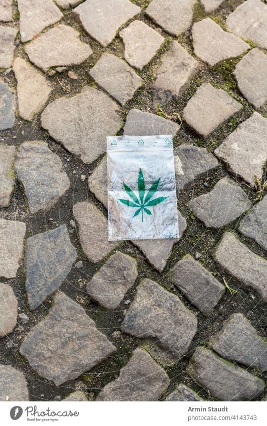 Cannabisblatt bedruckt auf einer kleinen Plastiktüte, die auf dem Bürgersteig liegt Sucht Hintergrund Tasche Blütenknospen Großstadt Kopfsteinpflaster