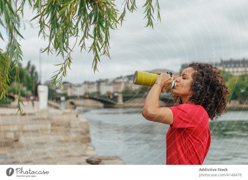 Durstige ethnische Sportlerin trinkt Wasser, nachdem sie in der Nähe eines Flusses trainiert hat trinken Flasche Headset Sportbekleidung Pause Baum benutzend