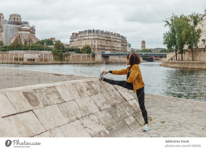 Ethnische Sportlerin mit Headset streckt ihr Bein in der Nähe eines Flusses in der Stadt Athlet Dehnung Drahtlos zuhören Musik Training benutzend Gerät Übung