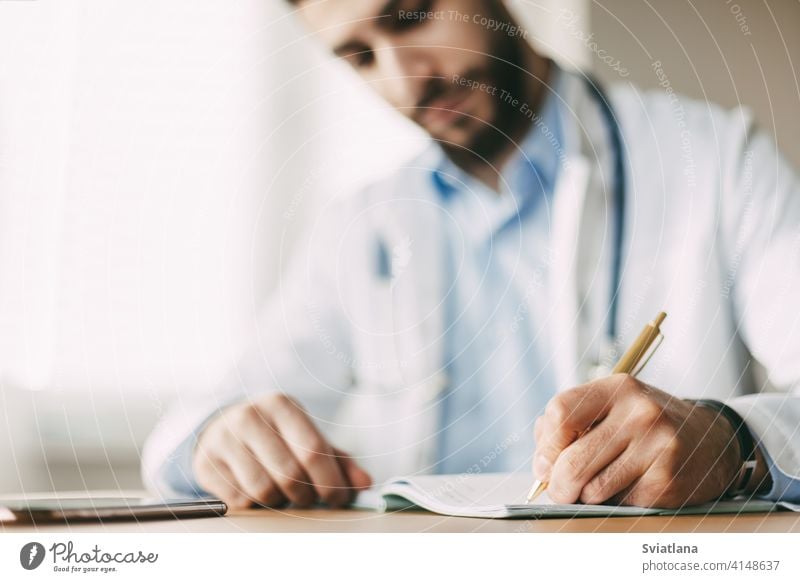 Freundlicher junger männlicher Arzt mit orientalischem Aussehen sitzt an einem Schreibtisch in seinem Büro und macht sich Notizen in einem Notizbuch, Nahaufnahme