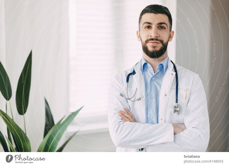 Lächelnder junger Arzt mit Stethoskopen und weißem Arztkittel steht mit verschränkten Armen in seinem Büro Medizin Mann Gesundheit medizinisch professionell