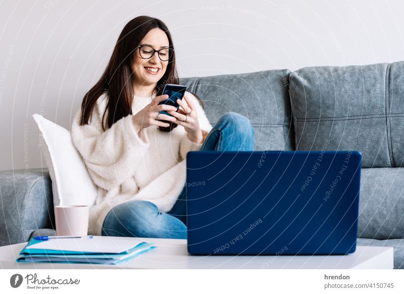 Frau im Gespräch mit Handy während der Arbeit mit Laptop sitzt auf einer Couch zu Hause im Innenbereich benutzend arbeiten Büro Job Notebook jung Glück Tippen