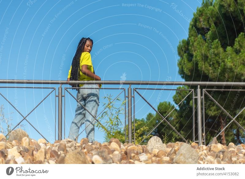 Stilvolle ethnische Frau steht in der Nähe Geländer im Park trendy Mode urban Hipster jung farbenfroh tausendjährig Geflecht Outfit schwarz lässig Jeanshose