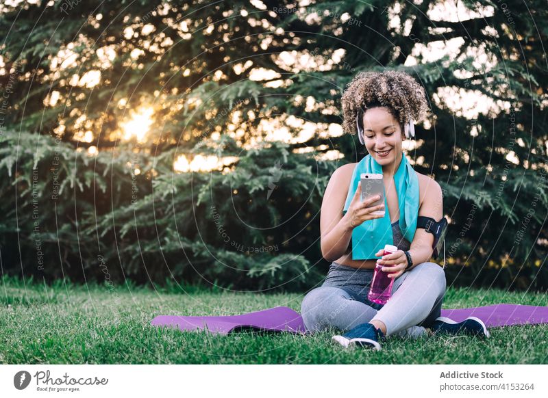 Sportliche Frau benutzt ihr Smartphone nach dem Training im Park sportlich benutzend ruhen heiter passen jung Browsen Kopfhörer Sommer Apparatur Fitness Gerät