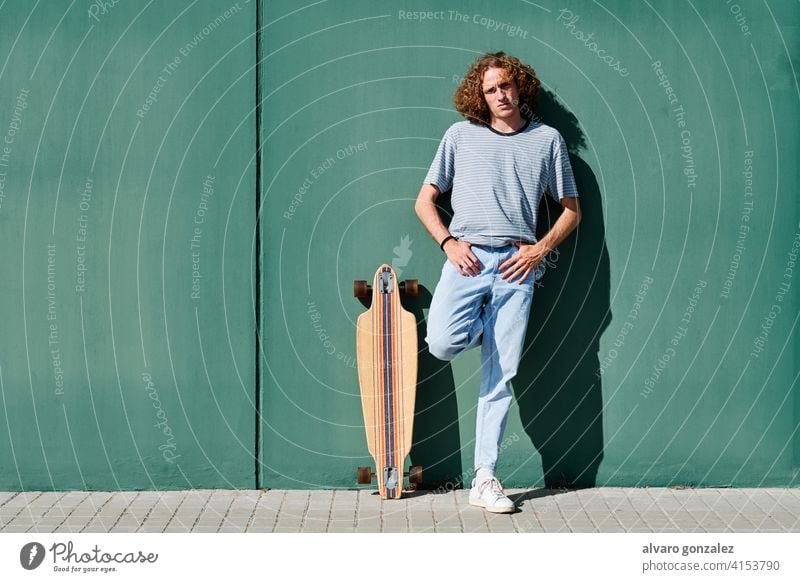 ein junger Mann mit lockigem Haar, der an einem sonnigen Tag mit seinem Skate- oder Longboard an einer grünen Wand steht che Skateboard attraktiv Person