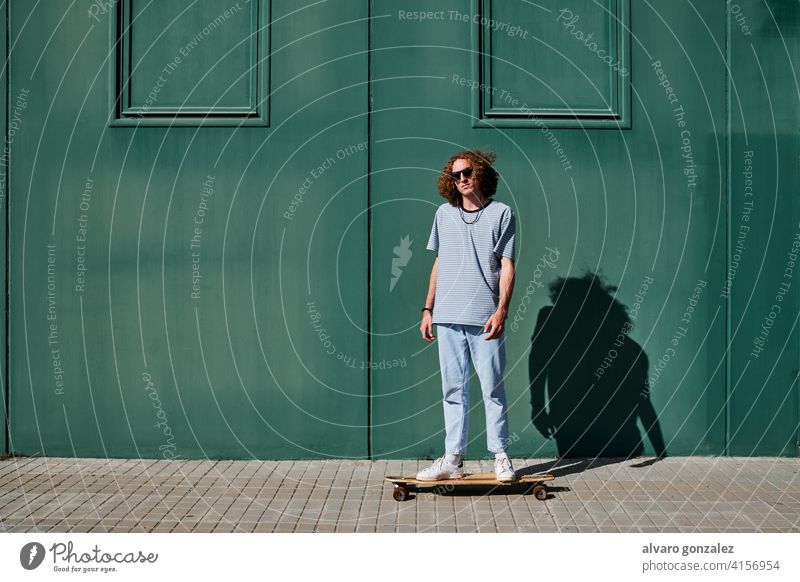 ein junger Mann mit lockigem Haar, der im Freien Schlittschuh läuft, mit einer grünen Wand hinter ihm Longboard che Skateboard attraktiv Person Skateboarding