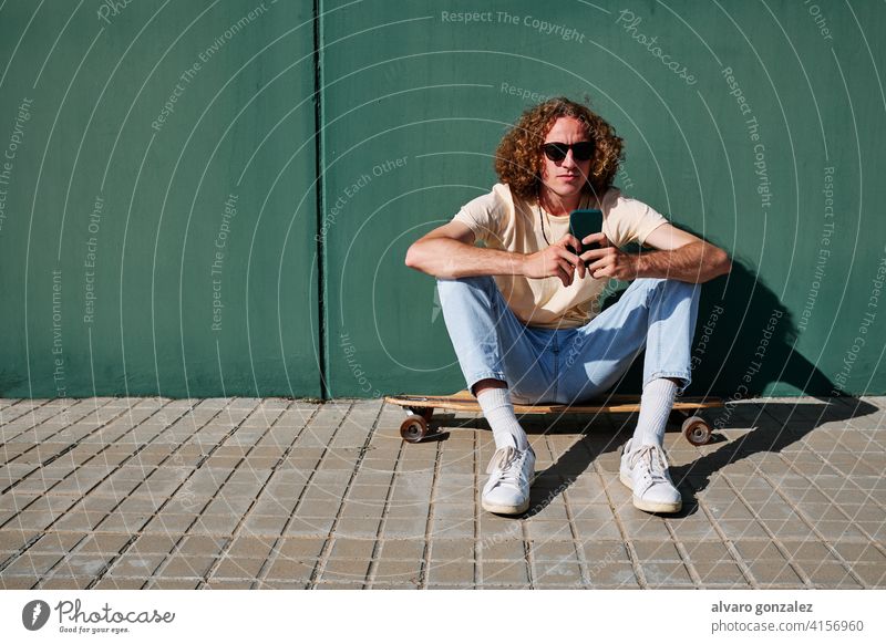ein junger Mann, der sein Smartphone benutzt und auf seinem Skateboard oder Longboard sitzt, mit einer Wand im Rücken che attraktiv Person Skateboarding Sport