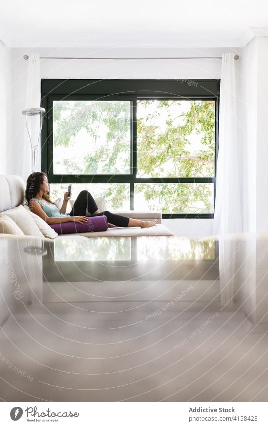 Ruhige Frau entspannt auf Sofa mit Smartphone und Yogamatte Browsen heimwärts Unterlage Lügen benutzend Wochenende sich[Akk] entspannen Sportbekleidung