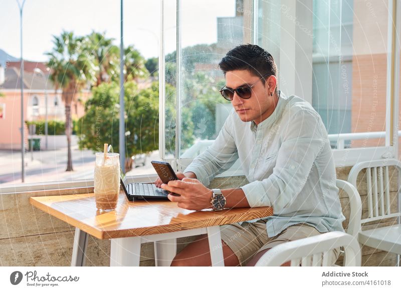 Konzentrierter junger Mann arbeitet aus der Ferne mit Laptop und Smartphone benutzend Arbeit freiberuflich Fokus online Anschluss Internet männlich Stil