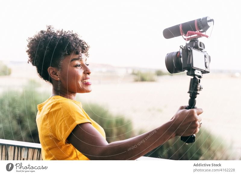 Positive Bloggerin dreht Video auf Terrasse vlog Selfie-Stick Fotokamera Aufzeichnen heiter reisen schießen Glück jung Frau Afroamerikaner schwarz ethnisch