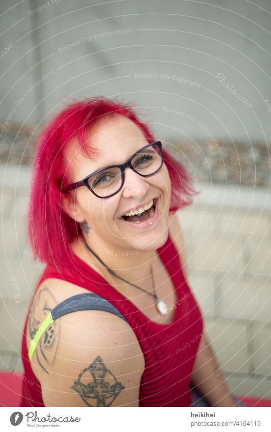 Eine junge rothaarige Frau mit Brille und Tattoos schaut lachend in die Kamera frau brille tattoos portrait lippenpiercing auffallend erscheinungsbild