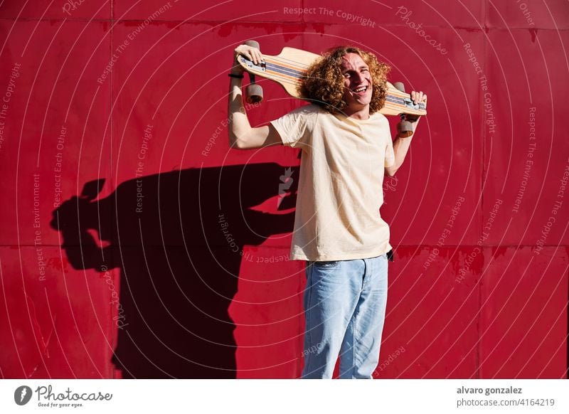 junger Mann mit lockigem Haar, der in die Kamera schaut, mit seinem Skateboard auf den Schultern und einem roten Hintergrund Longboard che attraktiv Person