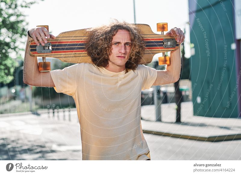junger Mann mit lockigem Haar, der mit seinem Longboard oder Skeateboard auf den Schultern mitten auf der Straße in die Kamera schaut che Skateboard attraktiv