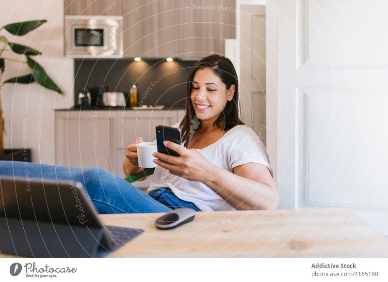 Smiley-Mädchen benutzt ein Smartphone in der Küche im Innenbereich heimwärts Haus Frau kaufen Kaffee Frühstück Morgen Kaukasier brünett Tisch Lifestyle lässig