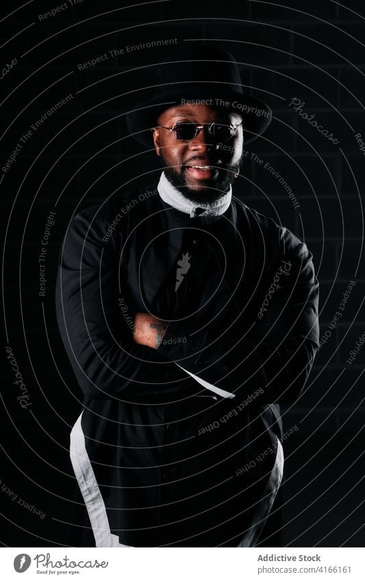 Ruhiger stilvoller Mann in dunklem Studio Stil schwarz Stoff Atelier Ring Hut Bekleidung trendy Sonnenbrille männlich ethnisch Afroamerikaner rund Form Lächeln