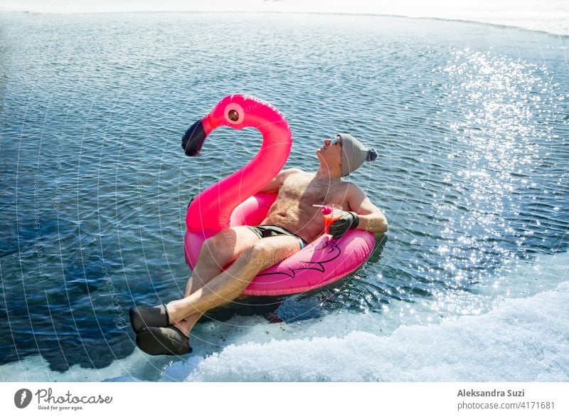 Ein Mann, der im Winter in Finnland in einem Eisloch schwimmt, schwebt auf einem rosa aufblasbaren Flamingo mit einem Cocktail in der Hand. Urlaubsoptionen, die vom Sommer träumen.