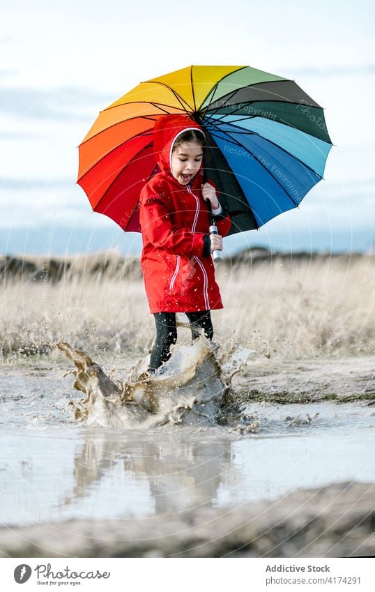 Fröhliches Mädchen mit Regenschirm spielt in einer Pfütze platschen Wasser Kind Zusammensein Kindheit Regenmantel Spaß haben spielerisch Gummi Stiefel spielen