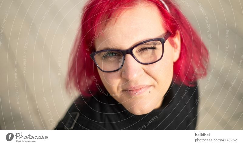 Eine rothaarige Frau mit Brille und Piercings schaut zwinkernd in die Kamera frau brille portrait lippenpiercing auffallend erscheinungsbild ausstrahlung jung
