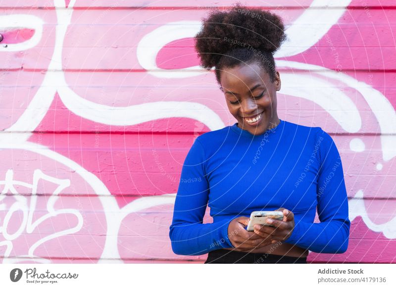 Zufriedene schwarze Frau, die in der Stadt auf ihrem Smartphone surft Browsen zuschauend Graffiti Wand Großstadt Straßenkunst heiter ethnisch Afroamerikaner