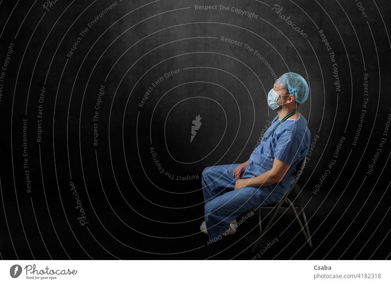 Trauriger, erschöpfter Arzt sitzt in einem dunklen Raum traurig müde Mann Seitenansicht Sitzen enttäuscht Auge zugeklappt Pandemie männlich Stress medizinisch
