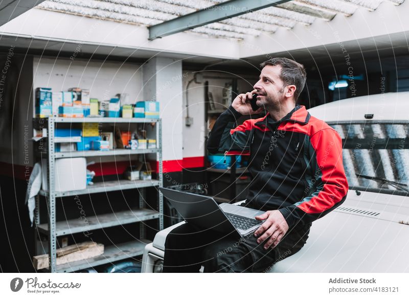 Automechaniker mit Laptop, der in der Werkstatt über sein Smartphone spricht Mechaniker sprechen Multimedia professionell Telefonanruf Mann Garage benutzend
