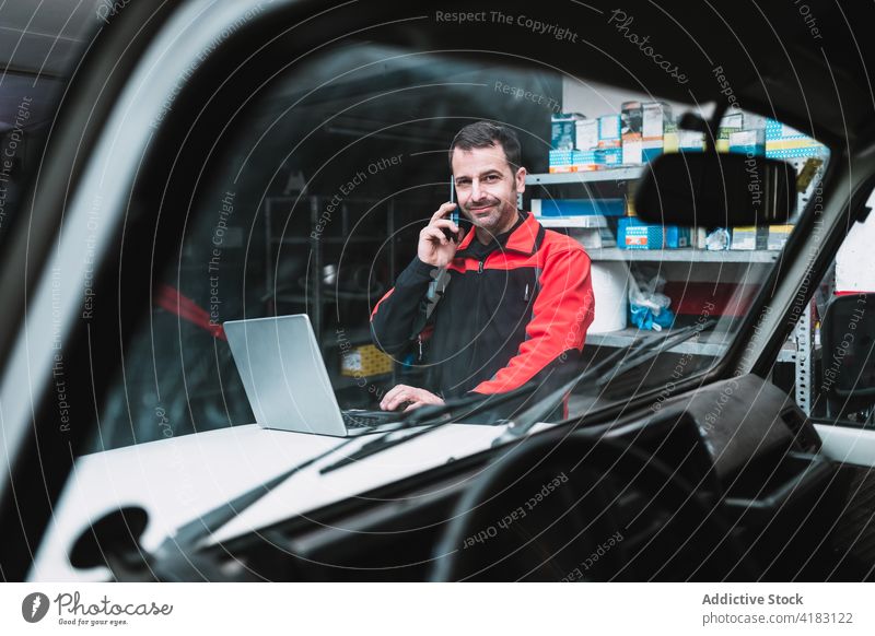 Automechaniker mit Laptop, der in der Werkstatt über sein Smartphone spricht Mechaniker sprechen Multimedia professionell Telefonanruf Mann Garage benutzend