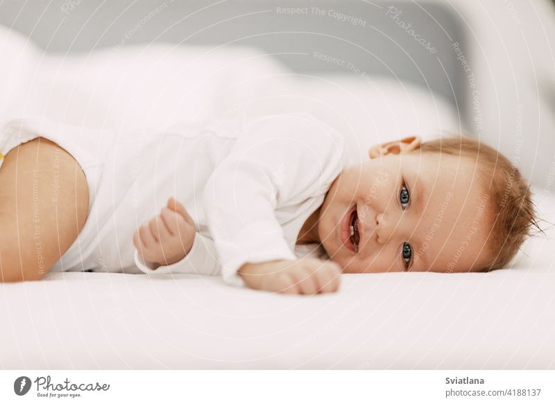 Ein blauäugiges Baby liegt in einem schneeweißen Bettchen unter einer Decke, lacht und schwelgt Familie Spaß Sitzen Spielzeug Kind Gesundheit heimwärts