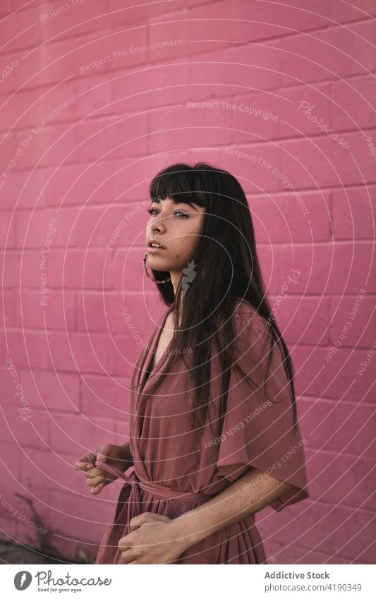 Modische junge ethnische Dame steht in der Nähe von rosa Wand und schaut weg Frau besinnlich nachdenklich Stil cool Mode Windstille Straße verträumt traumhaft