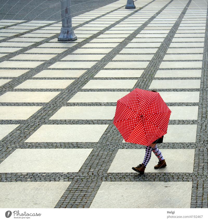 Frau mit bunten Strümpfen hinter einem rot-weiß gepunkteten Regenschirm auf einem großen Platz mit grafischer Pflasterung Mensch Beine Schuhe gemustert