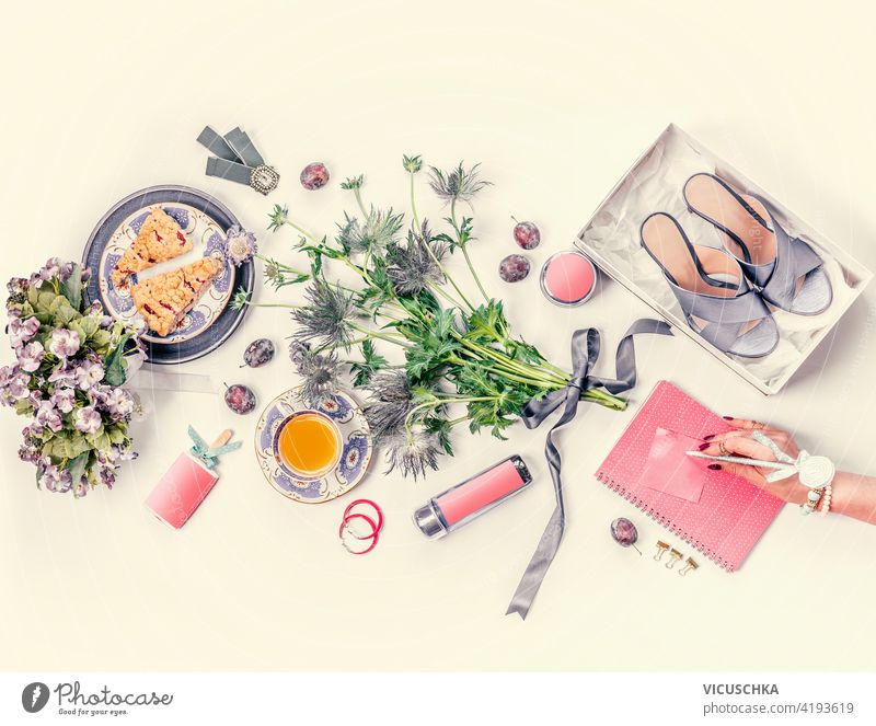 Frauen Hand mit rosa Notebook Lebensstil flach lag mit blauen neuen High Heels Schuhe in der Box, Blumenstrauß, Tasse Tee, Kuchen, Schmuck, Kosmetik, Früchte auf weißem Schreibtisch. Ansicht von oben. Schönheit und Mode