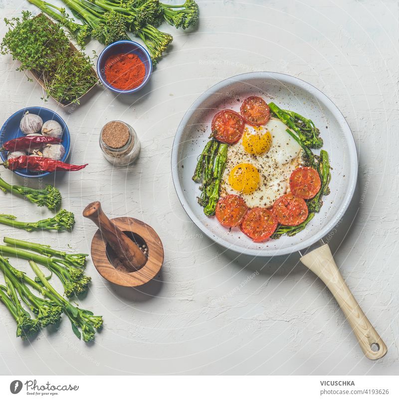Spiegeleier mit Tomaten und Brokkoli in einer weißen Bratpfanne auf einem Küchentisch mit Zutaten. Ansicht von oben. Gesundes Frühstück Tisch Draufsicht